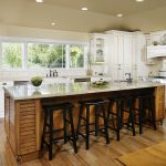 beige bambooo floring in kitchen with wooden island design before black wooden stools beneath small recessed ceiling lamps aside white cabinetry with some greenery