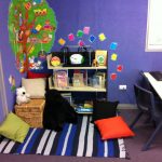 A reading nook for kids consisting a rattan box as bench plus pillows colorful rug with strip pattrern mini book shelves a computer desk with chair