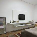 Small flat TV mounted on wall floating white TV console in two layers some decorative pieces a simple wooden chair a bed furniture bedroom rug