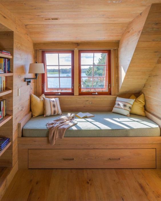 cozy reading nook fully covered in wood a couple of glass windows with red frame and trims bench seat with cushion addition and under drawer some throw pillows