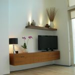 floating TV cabinet made from wood that is mounted in the corner a flat TV a table lamp a beautiful white vase with flower ornament a floating shelf for putting ceramic decorative vases wood flooring