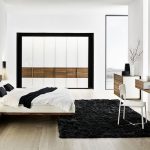 A modern bedroom with black wool area rug a modern vanity integrated with frameless mirror a white chair wooden drawer system lof bed with headboard plus bedside table modern wall lamps