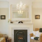 Square mirror with white concrete frame built over white fireplace a luxurious crystal pendant lighting fixture a rattan armchair with white wool pillows