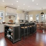 two-tier-Kitchen-Island-with-Side-Storage-in-black-with-light-granite-countertops-on-warm-hardwood-flooring-and-white-cabinetry-with-three-tall-pub-chairs