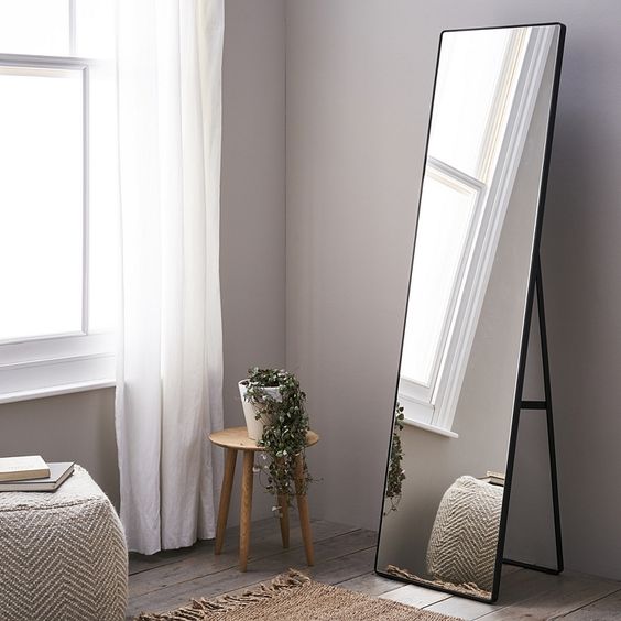 light wood stool with potted greenery free standing mirror with black wood finish frame flat woven runner white pouf chair whitewashed wood plank floors dramatically white draperies